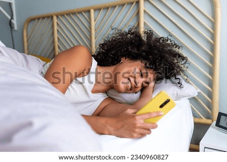 Happy woman using mobile phone in bed. Smiling black latina female looking mobile phone in the morning after waking up. Royalty-Free Stock Photo #2340960827