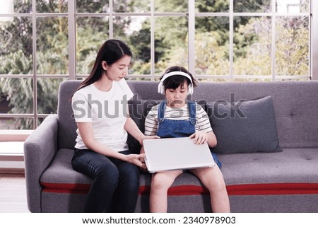 Smiling Asian mother and little girl child is relaxing with headphone and laptop for listening to online music or cartoon movie in social media on sofa in living room. Education and technology concept