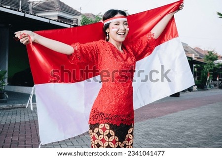 Happy smiling Indonesian woman wearing red kebaya holding Indonesia's flag to celebrate Indonesia Independence Day. Outdoor photoshoot concept