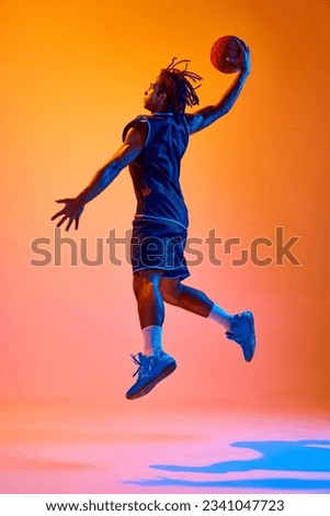 Full-length image of young sportsman, basketball player in motion, jumping with ball against orange background in neon lights. Concept of professional sport, competition, hobby, game, competition, ad Royalty-Free Stock Photo #2341047723
