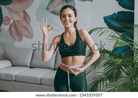 Young strong sporty athletic fitness trainer instructor woman wears green tracksuit hold measure tape on waist show ok okay training do exercises at home gym indoor. Workout sport motivation concept