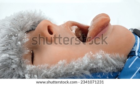 Portrait of boy lying in a snow-covered park, catching snowflakes with his tongue and smiling. Concept of outdoor winter play