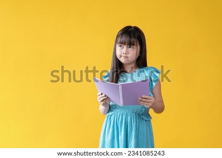 An adorable young Asian girl with a book in her hands is standing against an isolated yellow studio background with a thoughtful and doubtful facial expression. Kids, elementary school student