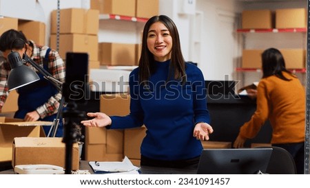Business owner filming teleshopping video on camera, advertising merchandise in storage room. Young woman using social media streaming platform to create online promotion, marketing. Tripod shot.