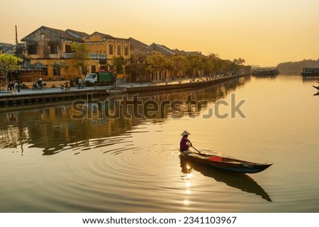 Awesome view of Vietnamese woman in traditional bamboo hat on wooden boat on the Thu Bon River at Hoi An Ancient Town at sunrise in Vietnam. Hoian is a popular tourist destination of Asia. Royalty-Free Stock Photo #2341103967