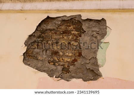 A closeup photo of a beige painted wall with a large area of peeled off paint and plaster in the center that shows ancient bricks bared.
