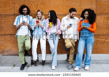 Diverse young confident people smiling and using their phones outside in a city while leaning against a wooden wall. Multiracial group of friends texting on their smartphones  Royalty-Free Stock Photo #2341150465
