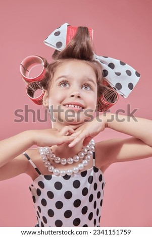 Fashion for kids - clothes and accessories. A little pretty girl in an elegant polka-dot dress and curlers on her head is going to a party and doing her make-up. Pink studio background. Pin-up style.