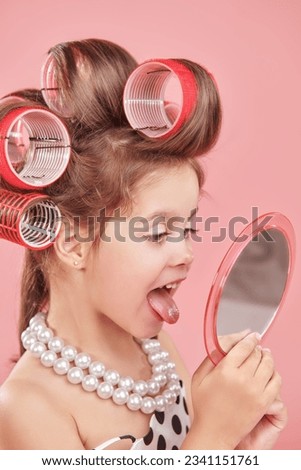 A little pretty girl in an elegant polka-dot dress and curlers on her head is going to a party and doing her make-up. Pink studio background. Kid's Fashion - clothes and accessories. Pin-up style.