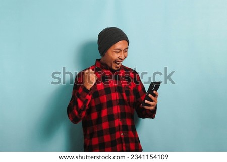 Excited Young Asian man with beanie hat and red plaid flannel shirt is making YES gesture with clenched fist while holding smartphone with good news, doing winner gesture, isolated on blue background Royalty-Free Stock Photo #2341154109
