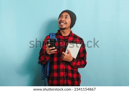 Excited young Asian male student, wearing backpack, beanie hat, and red plaid flannel shirt, looks aside at empty copy space, holds a book and uses his phone, while standing against a blue background