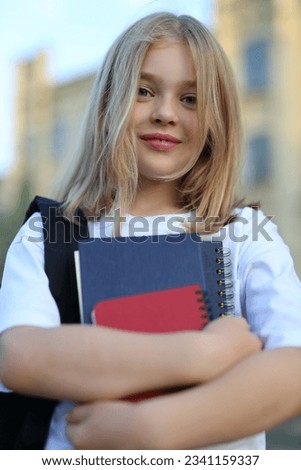 A school girl with a backpack and a notebook stands in front of a building on a sunny day.