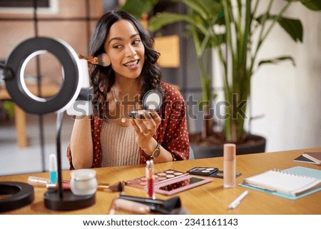 Young woman applying makeup on face with brush while recording vlog on smartphone for social media with LED ring lamp on table. Multiethnic fashion makeup artist vlogger live streaming makeup tutorial
