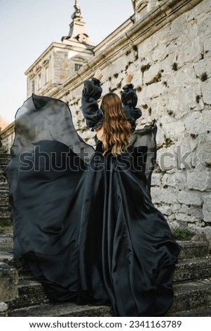 Woman in long black dress stands on stairs near ancient palace at sunset. Model girl in nature autumn day. Luxury female near old Pidhirtsi Castle, Lviv region, Ukraine. Stylish bride. Back view.