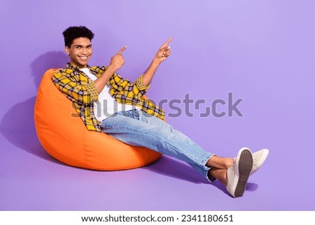 Full size portrait of cheerful nice person sitting comfy bag indicate fingers empty space isolated on purple color background