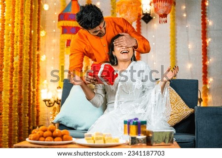 Happy brother by closing eyes giving surprise gift to sister during raksha bandhan festival celebration at home - concept of relationship bonding, togetherness and family ceremony.