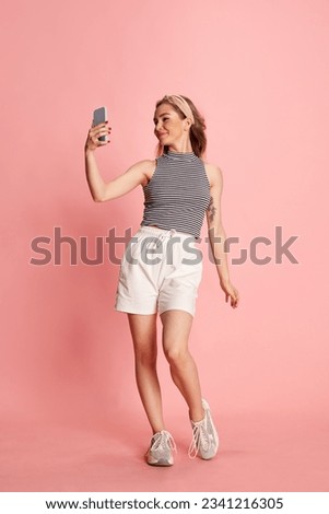 Full body photo of cheerful girl having video-call holding smart phone in hand over pink background. Concept of fashion, beauty, emotions. Ad