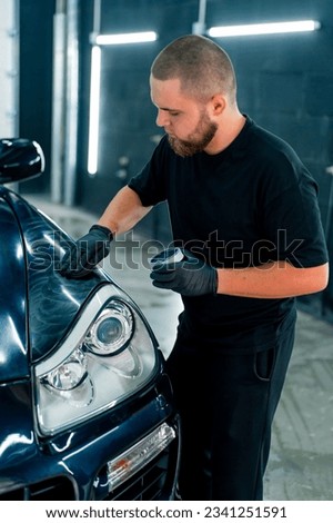 A focused car wash worker in black gloves polishes the hood and headlight of a luxury black car with car care concept polishing sponge