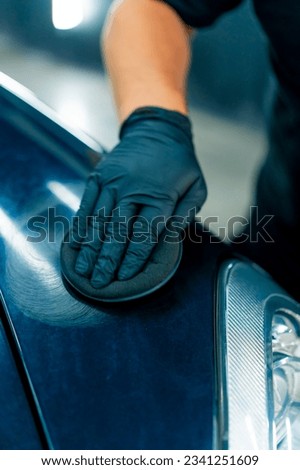 A close-up of a car wash worker in black gloves polishing the hood and headlight of a luxurious black car with a car polishing sponge