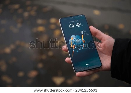 Woman checking weather using app on smartphone on street, closeup. Data and illustration of cloud with lighting and rain on screen Royalty-Free Stock Photo #2341256691