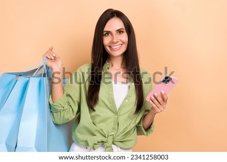 Photo of cheerful adorable girl with straight hairstyle dressed green blouse hold bags smartphone isolated on beige color background