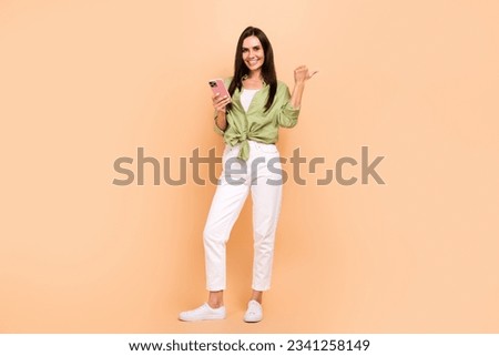 Full length photo of optimistic woman wear green shirt indicating at eshop empty space hold smartphone isolated on beige color background