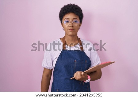 Young african american woman wearing professional waitress apron holding clipboard looking sleepy and tired, exhausted for fatigue and hangover, lazy eyes in the morning. 