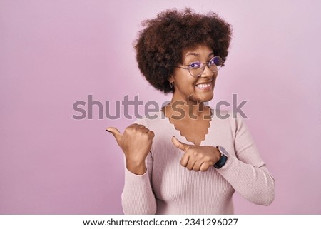 Young african american woman standing over pink background pointing to the back behind with hand and thumbs up, smiling confident 