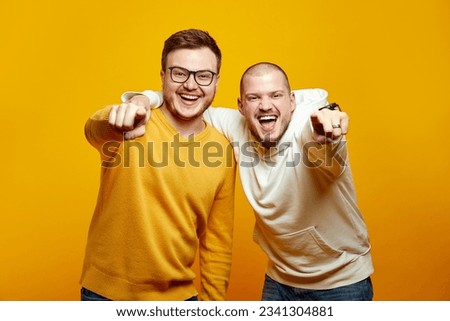 Portrait of two handsome happy young men embracing each other and pointing fingers at camera while smiling, standing isolated over yellow orange studio background Royalty-Free Stock Photo #2341304881
