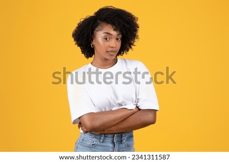 Serious confident young black curly woman in white t-shirt with crossed arms, thinking, isolated on orange background, studio. Ad and offer, human emotions, work and study