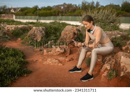Glad millennial caucasian lady athlete in sportswear, wireless headphones chatting on smartphone, rest from workout, sits on stone, full length. Sports, app, body care and social network outdoor