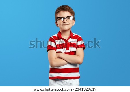 Positive preschool kid boy in red striped shirt and nerdy eyeglasses crossing arms and looking at camera against bright blue background