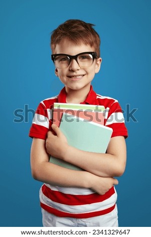 Vertical photo of little genius kid in nerdy glasses and red striped shirt holding notebook while standing against blue background during studies.