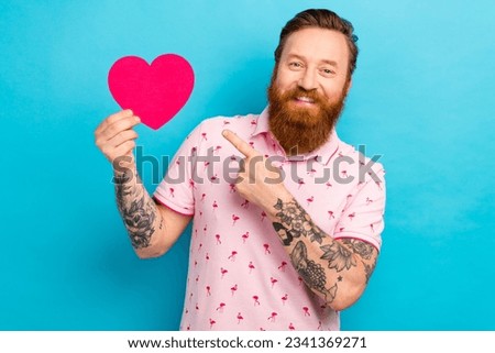 Photo of candid sincere man with redhair beard wear pink t-shirt directing at big shape heart postcard isolated on blue color background