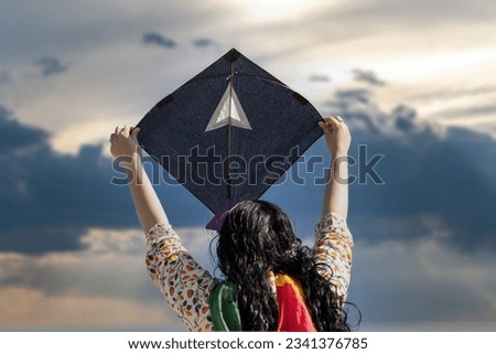 Young girl in traditional indian clothing holding black kite high above head launching it on sankranti republic independence day celebrations Royalty-Free Stock Photo #2341376785