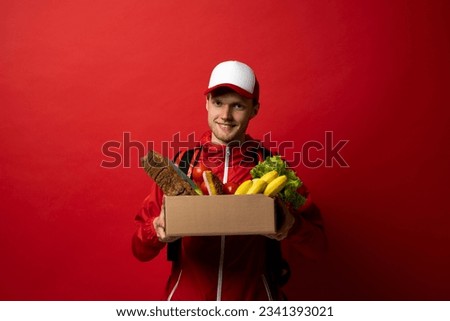Deliver man worker in red uniform handling box of food, fruit, vegetable. Postman and express grocery delivery service concept. Royalty-Free Stock Photo #2341393021