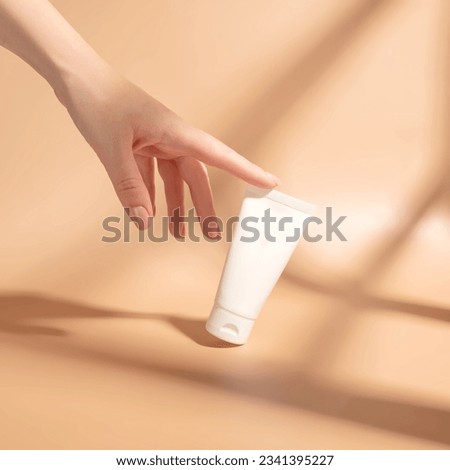 Woman's hand touches a mockup of white tube of cream. Unlabeled packaging for cosmetics in sunlight on beige background. Concept of skincare. Harsh shadows Royalty-Free Stock Photo #2341395227