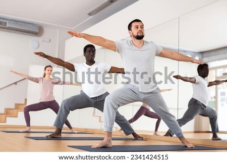 Caucasian man practising warrior II pose with people during group yoga training. Royalty-Free Stock Photo #2341424515