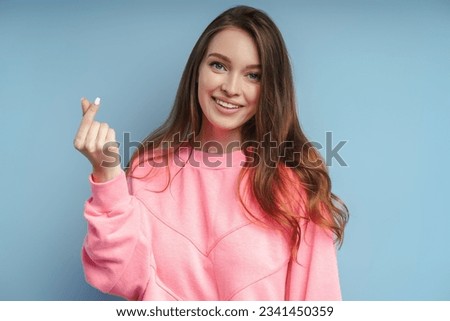 Portrait of smiling woman with long hair in pink sweatshirt gesturing showing heart from fingers, k pop culture, isolated on blue background. Young modern female with toothy smile looks at camera
