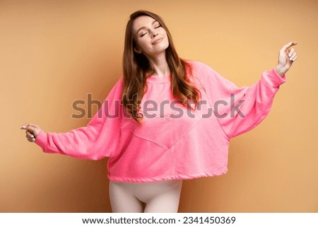 Portrait of beautiful smiling Caucasian woman with closed eyes wearing pink sweatshirt dancing isolated on beige background. Attractive female taking her favorite hobby. Positive lifestyle concept