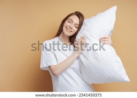 Beautiful smiling woman with closed eyes wearing white t shirt holding pillow isolated on beige background. Cute female posing. Concept of shopping, store, advertisement Royalty-Free Stock Photo #2341450375