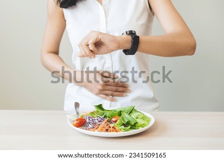 Intermittent fasting, health care asian young woman dietary, having stomach ache, temptation hungry of breakfast food in morning on table at home, looking at watch on her wrist but not yet time to eat Royalty-Free Stock Photo #2341509165