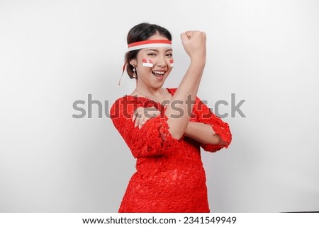 Excited Asian woman wearing a red kebaya and headband showing strong gesture by lifting her arms and muscles smiling proudly. Indonesia's independence day concept.
