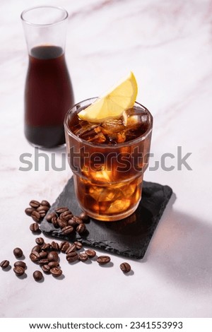 Cold brew coffee in a glass with ice and lemon on a light marble background with coffee beans and bottle. Concept summer craft refreshing homemade drink. Free space for text.