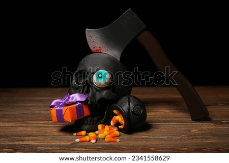 Composition with beautiful gift box, tasty candy corns and ax in skull for Halloween on wooden table against dark background, closeup