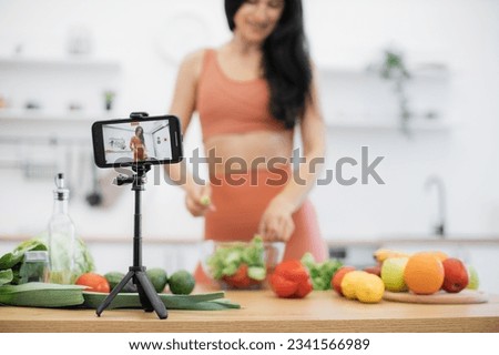 Focus on handheld device on tripod shooting attractive lady putting fresh greens into salad at kitchen table. Young fitness instructor filming virtual lesson on dishes rich in vitamins and nutrients.