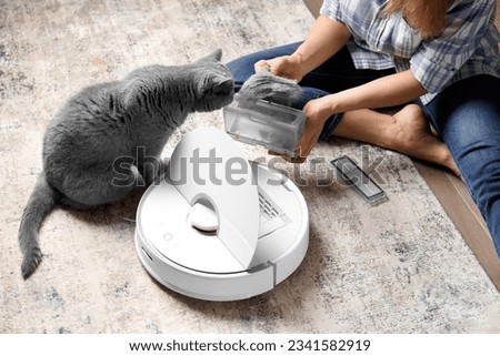 The container of the vacuum cleaner robot is in female hands, the dust collector of the robot vacuum cleaner is full of wool, the cat sits nearby and looks at the container. Royalty-Free Stock Photo #2341582919