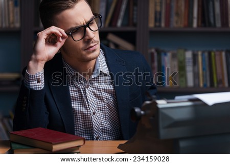 Thoughtful author. Thoughtful young author working at the typewriter and adjusting his eyeglasses while sitting at his working place with bookshelf in the background 