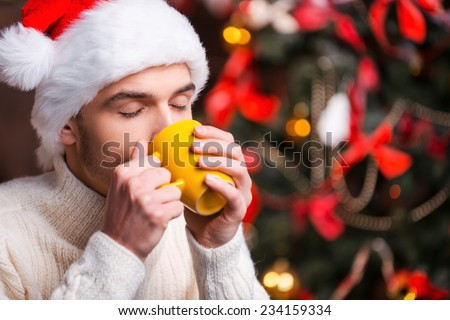 Hot drink at winter night. Handsome young man in Santa hat drinking hot drink with Christmas Tree in the background 