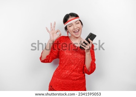 A cheerful Asian woman wearing red kebaya and headband, holding her phone while gesturing OK sign with her fingers, isolated by white background. Indonesia's independence day concept